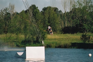 A person is wakeboarding in a body of water, being pulled by a cable system. The wakeboarder is performing a jump or trick above the water, with a lush, green forest and grassy area in the background. A white ramp or obstacle is visible in the foreground, and the sky is clear.