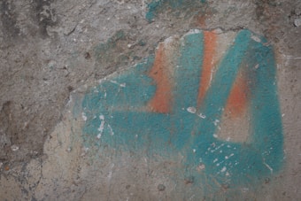 A textured concrete surface with weathered blue and orange paint, showing signs of aging and chipping. The colors are irregularly distributed over the rough, uneven surface.