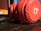 A close-up view of a pair of red headphones resting on a dark wooden surface next to a laptop. The headphones are prominently displayed with a focus on the circular ear cup featuring a recognizable logo.