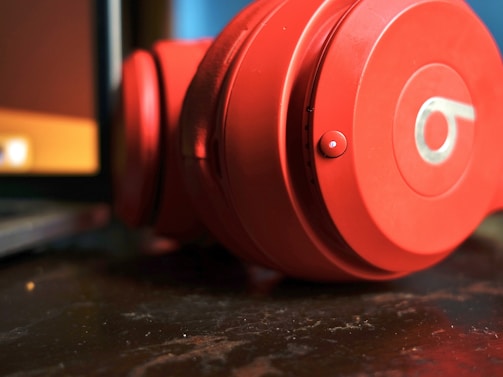 A close-up view of a pair of red headphones resting on a dark wooden surface next to a laptop. The headphones are prominently displayed with a focus on the circular ear cup featuring a recognizable logo.
