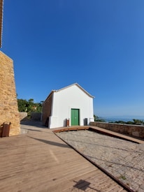 A small, white building with a green door is situated on a paved area. The building blends rustic and modern elements, with stone walls adjacent to it. A clear blue sky dominates the background, and some greenery is visible in the distance alongside a glimpse of the ocean.