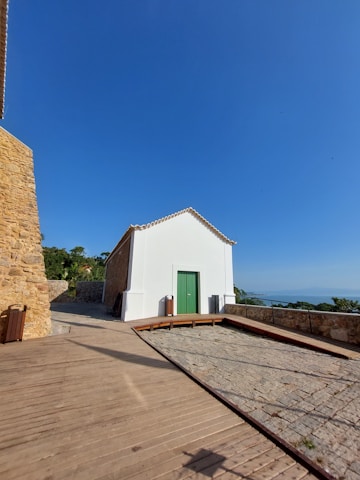 A small, white building with a green door is situated on a paved area. The building blends rustic and modern elements, with stone walls adjacent to it. A clear blue sky dominates the background, and some greenery is visible in the distance alongside a glimpse of the ocean.