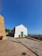 A small, white building with a green door is situated on a paved area. The building blends rustic and modern elements, with stone walls adjacent to it. A clear blue sky dominates the background, and some greenery is visible in the distance alongside a glimpse of the ocean.