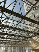 A complex metal framework creating a geometric pattern against a bright sky. Glass panels in the roof allow natural light to illuminate the interior, casting intricate shadows on the surrounding surfaces.