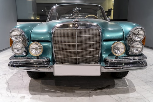A vintage car with a polished teal exterior features a prominent grille and dual round headlights on each side. The vehicle is showcased in a well-lit indoor setting with a reflective floor surface.