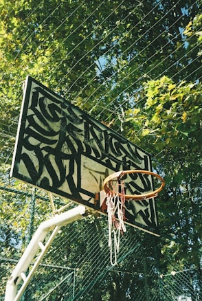 An outdoor basketball hoop features a uniquely designed backboard with an abstract black and white pattern. The hoop is slightly rusty, and the net is worn and tangled. There are tall, lush green trees in the background, with sunlight filtering through the leaves, giving the scene a natural and serene setting.