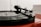 A close-up view of a vinyl record player with a spinning black record. The tonearm, with a 'SUMIKO' cartridge, is placed on the record. The turntable sits on a red surface with a blurred white background.