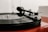 A close-up view of a vinyl record player with a spinning black record. The tonearm, with a 'SUMIKO' cartridge, is placed on the record. The turntable sits on a red surface with a blurred white background.
