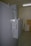 A large gray industrial electrical panel with an on/off switch is mounted on the side of a gray wall. Above the panel are multiple electrical conduits running upward. The surrounding area appears to be a utility or storage room with a wooden board and some equipment in the background.