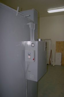 A large gray industrial electrical panel with an on/off switch is mounted on the side of a gray wall. Above the panel are multiple electrical conduits running upward. The surrounding area appears to be a utility or storage room with a wooden board and some equipment in the background.