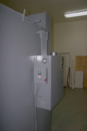 A large gray industrial electrical panel with an on/off switch is mounted on the side of a gray wall. Above the panel are multiple electrical conduits running upward. The surrounding area appears to be a utility or storage room with a wooden board and some equipment in the background.