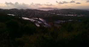 Solar panels spread across a hillside overlooking a coastal city at dusk, with a view of the ocean and distant islands on the horizon. The landscape is illuminated by the soft glow of the setting sun, casting warm hues over the clouds and water.