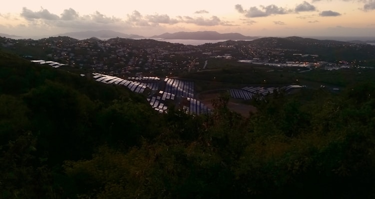 Solar panels spread across a hillside overlooking a coastal city at dusk, with a view of the ocean and distant islands on the horizon. The landscape is illuminated by the soft glow of the setting sun, casting warm hues over the clouds and water.
