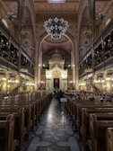 An ornate interior of a large synagogue featuring a high arched ceiling with intricate designs and a grand chandelier. Wooden pews line both sides leading towards an elaborate, gold-adorned altar. People are seated on the right side, and a small flag featuring the Union Jack is visible along the center aisle.