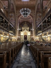 An ornate interior of a large synagogue featuring a high arched ceiling with intricate designs and a grand chandelier. Wooden pews line both sides leading towards an elaborate, gold-adorned altar. People are seated on the right side, and a small flag featuring the Union Jack is visible along the center aisle.