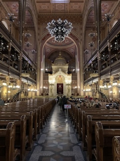 An ornate interior of a large synagogue featuring a high arched ceiling with intricate designs and a grand chandelier. Wooden pews line both sides leading towards an elaborate, gold-adorned altar. People are seated on the right side, and a small flag featuring the Union Jack is visible along the center aisle.