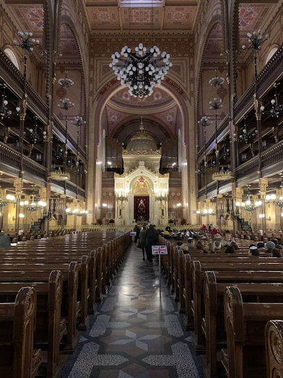 An ornate interior of a large synagogue featuring a high arched ceiling with intricate designs and a grand chandelier. Wooden pews line both sides leading towards an elaborate, gold-adorned altar. People are seated on the right side, and a small flag featuring the Union Jack is visible along the center aisle.