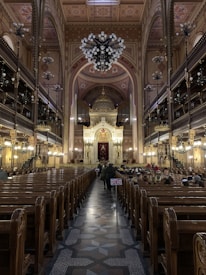 An ornate interior of a large synagogue featuring a high arched ceiling with intricate designs and a grand chandelier. Wooden pews line both sides leading towards an elaborate, gold-adorned altar. People are seated on the right side, and a small flag featuring the Union Jack is visible along the center aisle.