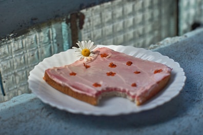 A slice of bread with pink frosting on a white plate. Small orange flowers are sprinkled on top of the frosting. A white daisy with a yellow center is placed on the bread as a decorative element. The setting appears to be a rustic environment with a textured surface and windows in the background.