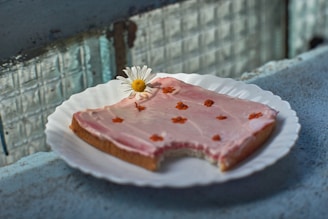 A slice of bread with pink frosting on a white plate. Small orange flowers are sprinkled on top of the frosting. A white daisy with a yellow center is placed on the bread as a decorative element. The setting appears to be a rustic environment with a textured surface and windows in the background.