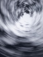 A swirling, abstract vortex of blurred lines creates a dynamic circular pattern. The design is monochromatic, featuring shades of gray and white, which enhances the sense of motion and depth.