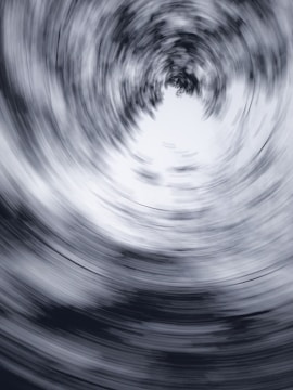 A swirling, abstract vortex of blurred lines creates a dynamic circular pattern. The design is monochromatic, featuring shades of gray and white, which enhances the sense of motion and depth.