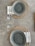 A neatly arranged table setting with two woven placemats, each featuring a gray ceramic bowl atop a decorative napkin with a leaf pattern. Accompanying the bowls are sets of silver cutlery and two clear wine glasses placed beside them. The background is a smooth, gray concrete surface.