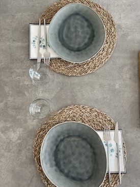 A neatly arranged table setting with two woven placemats, each featuring a gray ceramic bowl atop a decorative napkin with a leaf pattern. Accompanying the bowls are sets of silver cutlery and two clear wine glasses placed beside them. The background is a smooth, gray concrete surface.