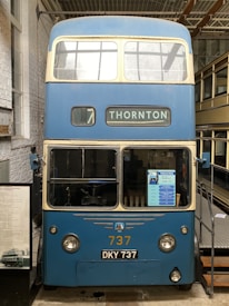 A vintage double-decker blue bus is displayed indoors. The bus has two levels with large windows and 'THORNTON' displayed at the front. Its number is 737 with a license plate DKY 737. The bus is showcased with informational signs nearby.