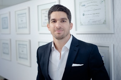 A man in a dark suit and white shirt stands in an office setting, smiling slightly. The background features a wall with framed certificates, indicating a professional environment.
