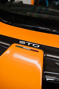 A close-up of a car's hood displaying the letters 'STO' and an Italian flag emblem. The surface is primarily orange with glossy black trim.