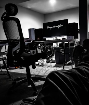 A dimly lit room with a modern workspace featuring a comfortable mesh office chair in front of a desk. On the desk, a computer monitor with the text 'Every Atom Of Me' is flanked by speakers and a PC tower. Cables and wires are scattered on the floor. The lower half of a person's legs is visible in the foreground, suggesting a relaxed posture.