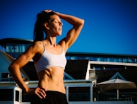 A fit woman in a sleeveless top and shorts stands outdoors with her hand resting on her head. Her expression is relaxed and contemplative as she looks upwards. The modern building and clear blue sky in the background suggest a warm, sunny day.