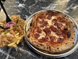 A close-up view of a pepperoni pizza on a metal plate beside a serving of fried food consisting of French fries topped with fried chicken, pickles, and onions. The items are placed on a marble or granite tabletop.