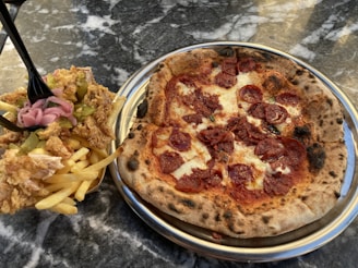 A close-up view of a pepperoni pizza on a metal plate beside a serving of fried food consisting of French fries topped with fried chicken, pickles, and onions. The items are placed on a marble or granite tabletop.