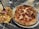 A close-up view of a pepperoni pizza on a metal plate beside a serving of fried food consisting of French fries topped with fried chicken, pickles, and onions. The items are placed on a marble or granite tabletop.