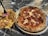 A close-up view of a pepperoni pizza on a metal plate beside a serving of fried food consisting of French fries topped with fried chicken, pickles, and onions. The items are placed on a marble or granite tabletop.