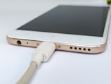 A smartphone is connected to a charger cable. The focus is on the bottom edge of the phone, showing the charging port, headphone jack, and speaker holes. The phone has a white front, with a metallic gold trim on the sides.