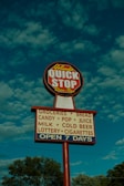 A tall sign with a round red and white top section labeled 'Yolanda QUICK STOP Drive-Thru' and a rectangular lower section listing items like groceries, bread, candy, pop, juice, milk, cold beer, lottery, and cigarettes. The sign also indicates it's open seven days a week. It is set against a blue sky with fluffy clouds and framed by trees at the bottom.