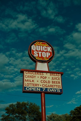 A tall sign with a round red and white top section labeled 'Yolanda QUICK STOP Drive-Thru' and a rectangular lower section listing items like groceries, bread, candy, pop, juice, milk, cold beer, lottery, and cigarettes. The sign also indicates it's open seven days a week. It is set against a blue sky with fluffy clouds and framed by trees at the bottom.