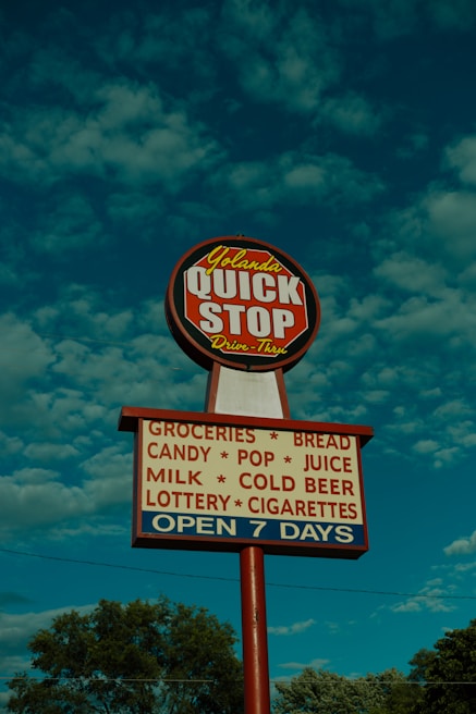 A tall sign with a round red and white top section labeled 'Yolanda QUICK STOP Drive-Thru' and a rectangular lower section listing items like groceries, bread, candy, pop, juice, milk, cold beer, lottery, and cigarettes. The sign also indicates it's open seven days a week. It is set against a blue sky with fluffy clouds and framed by trees at the bottom.
