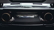 A close-up view of the rear end of a high-performance car, showcasing the McLaren logo prominently in the center. The design features sleek lines, a mesh grille, and dual exhaust pipes that suggest power and speed.
