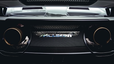 A close-up view of the rear end of a high-performance car, showcasing the McLaren logo prominently in the center. The design features sleek lines, a mesh grille, and dual exhaust pipes that suggest power and speed.