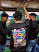 Three men are standing inside a moving vehicle, possibly a train or a bus. Each of them is holding onto metal handles for support. The man in the center is wearing a black shirt with a colorful, graphic design on the back, featuring various doodles and the words 'GO WEIRDO.' The other two men are facing each other, engaged in conversation.