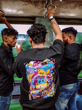 Three men are standing inside a moving vehicle, possibly a train or a bus. Each of them is holding onto metal handles for support. The man in the center is wearing a black shirt with a colorful, graphic design on the back, featuring various doodles and the words 'GO WEIRDO.' The other two men are facing each other, engaged in conversation.