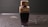 A perfume bottle with a sleek design featuring a dark lower half and a gold-colored cap sits on a wet surface. Water droplets are scattered around, enhancing the reflective appearance of the surface. The label on the bottle is embossed and includes text.