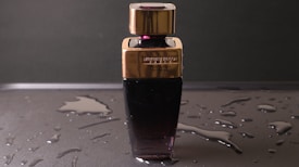 A perfume bottle with a sleek design featuring a dark lower half and a gold-colored cap sits on a wet surface. Water droplets are scattered around, enhancing the reflective appearance of the surface. The label on the bottle is embossed and includes text.