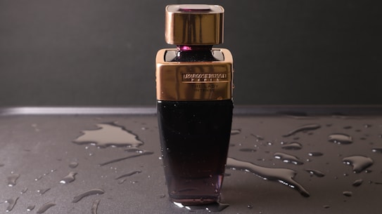 A perfume bottle with a sleek design featuring a dark lower half and a gold-colored cap sits on a wet surface. Water droplets are scattered around, enhancing the reflective appearance of the surface. The label on the bottle is embossed and includes text.