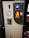A bottle recycling machine with a slot for inserting bottles, a circular sign indicating recycling, and a lit display. An information sheet with text in a foreign language is attached to the front.