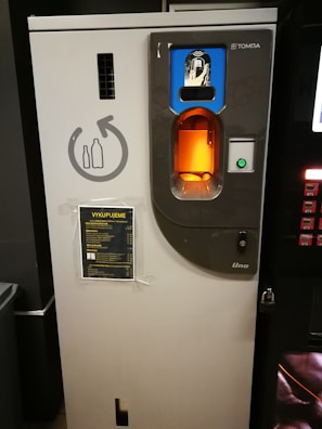 A bottle recycling machine with a slot for inserting bottles, a circular sign indicating recycling, and a lit display. An information sheet with text in a foreign language is attached to the front.
